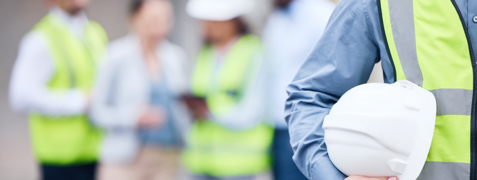 Worker holding a safety helmet with a team in safety vests in the background, representing workplace safety.